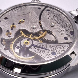 May include: Close-up of a silver-toned wristwatch with a black leather band. The watch's intricate mechanism is visible through the clear back, showcasing gears, jewels, and engraved details. The watch face displays the text "American Watch Co."