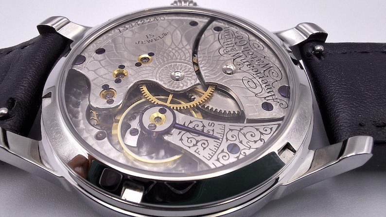 May include: Close-up of a silver-toned wristwatch with a black leather band. The watch's intricate mechanism is visible through the clear back, showcasing gears, jewels, and engraved details. The watch face displays the text "American Watch Co."