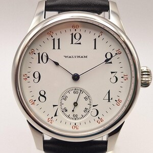 May include: A silver-toned Waltham wristwatch with a white face and black leather band. The watch face features black Arabic numerals, a subdial, and the brand name. Red minute markers encircle the face.