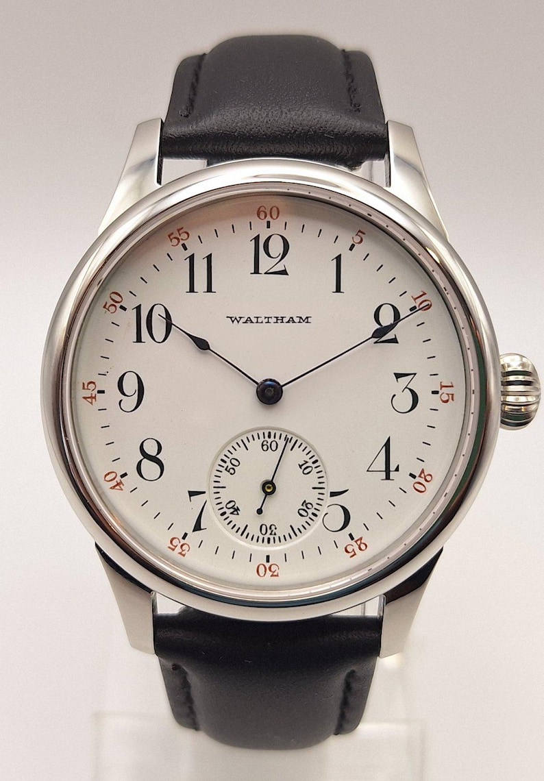 May include: A silver-toned Waltham wristwatch with a white face and black leather band. The watch face features black Arabic numerals, a subdial, and the brand name. Red minute markers encircle the face.