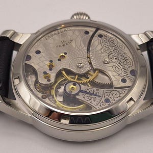 May include: Close-up of a silver-toned wristwatch with a black leather band. The watch's back reveals intricate mechanical components, including gears, jewels, and the text "American Watch Co." and "15 Jewels". The watch has a classic, vintage aesthetic.