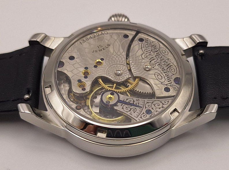 May include: Close-up of a silver-toned wristwatch with a black leather band. The watch's back reveals intricate mechanical components, including gears, jewels, and the text "American Watch Co." and "15 Jewels". The watch has a classic, vintage aesthetic.