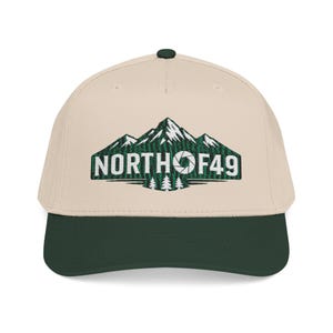 Könnte beinhalten: Eine beige Baseballkappe mit dunkelgrünem Schirm und einem Knopf oben. Die Vorderseite zeigt eine gestickte Grafik aus grünen Bergen und Tannen mit dem weißen Text NORTH OF 49, wobei das O durch ein Kamera-Blenden-Symbol ersetzt ist.