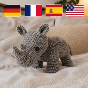 Puede incluir: Un juguete de rinoceronte de ganchillo gris con un cuerno prominente, orejas pequeñas y ojos de botón negros. El animal hecho a mano se exhibe sobre una superficie blanca, con banderas de Alemania, Francia, España y Estados Unidos en el fondo.