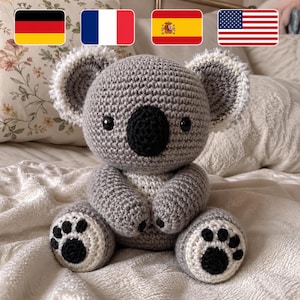 Puede incluir: Un koala de ganchillo hecho a mano en tonos grises, blancos y negros. El koala tiene una nariz redonda negra, ojos negros y orejas grandes y esponjosas. Las patas del oso tienen almohadillas negras. El koala está sentado sobre una superficie blanca.
