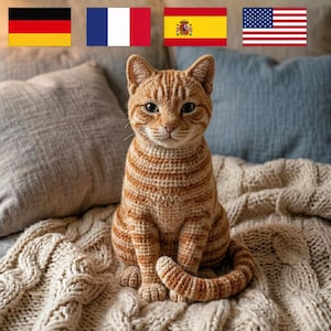 Op de afbeelding: Een gehaakte oranje tabby kattenfiguur met bruine strepen, zittend op een crèmekleurige gebreide deken. De kat heeft gedetailleerde kenmerken en is omgeven door kussens. Vlaggen van Duitsland, Frankrijk, Spanje en de VS op de achtergrond.