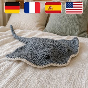 May include: A handmade, gray crochet stingray plush toy with a white border and black eyes. The plush toy is lying on a white textured surface. The image also shows the flags of Germany, France, Spain, and the United States.