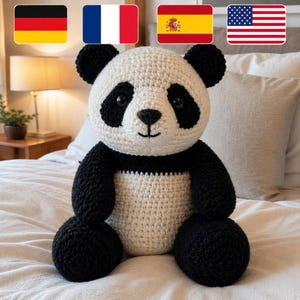 May include: A crocheted panda bear, featuring a white body with black arms, legs, and ears, and black markings around the eyes. The panda is sitting upright with a smiling face. The toy is handmade and placed on a bed with a neutral background.