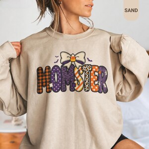 Puede incluir: Una sudadera de cuello redondo color arena con el texto MOMSTER en letras grandes con varios patrones, incluyendo cuadros, lunares, estampado de leopardo y rayas. Sobre el texto hay un lazo grande con un caramelo de maíz en el centro y murciélagos.