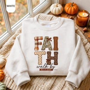 Puede incluir: Una sudadera blanca de cuello redondo con la palabra FAITH en letras grandes estampadas con lazos marrones. Debajo, el texto dice walk by NOT BY SIGHT 2 COR 5:7. Las letras presentan patrones de cuadros, flores, punto trenzado, lunares y leopardo.