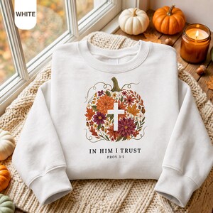 Puede incluir: Una sudadera blanca de cuello redondo con un gráfico central de una calabaza compuesta por flores y hojas de colores otoñales con una cruz blanca en el centro. Debajo del gráfico, el texto dice IN HIM I TRUST PROV 3:5 en letras negras.
