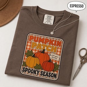 Puede incluir: Una camiseta color espresso con un gráfico de calabazas naranjas, hojas verdes y un fondo a cuadros. El texto dice PUMPKIN PATCH, SPOOKY SEASON y pick your favorite. La camiseta tiene una etiqueta Comfort Colors en el cuello.