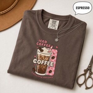 May include: A dark brown t-shirt featuring a graphic of an iced coffee drink with the text ICED COFFEE, COFFEE, and CUTE LITTLE CAFE DATE. The design includes a pink checkered background, coffee beans, and small hearts. A silver necklace rests on the shirt.