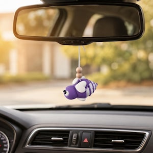 Könnte beinhalten: Ein lila bienenförmiges Auto-Accessoire, das an einem Rückspiegel an einer weißen Schnur mit zwei kleinen Holzperlen hängt. Die Biene hat weiße Flügel, einen gestreiften Körper und zwei kleine schwarze Augen und hängt im Fahrzeuginnenraum.
