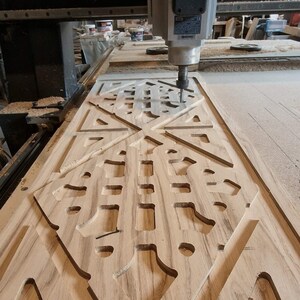 May include: A CNC machine with a silver spindle labeled AREL is carving a complex geometric pattern into a light brown wooden panel. The wood features visible grain lines and deep, precision-cut channels forming a symmetrical design.