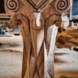 May include: A light brown wooden furniture leg featuring intricate hand-carved details. The top section displays a classic scroll-like volute design with geometric patterns, while the lower shaft has a faceted, diamond-shaped texture.