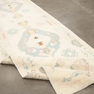 Könnte beinhalten: Cremefarbener Läufer mit einem geometrischen Muster in Blau-, Braun- und Pfirsichtönen. Der Teppich ist teilweise aufgerollt und zeigt sein kompliziertes Design und seine Textur. Der Teppich liegt auf einer grauen Oberfläche.