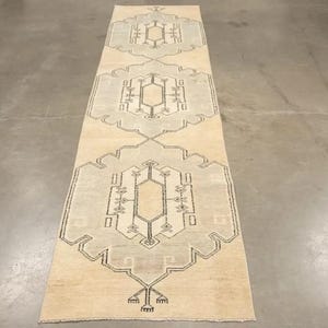 Könnte beinhalten: Ein rechteckiger, beigefarbener Läufer mit geometrischen Designs in Grau und Schwarz. Der Teppich weist sich wiederholende Muster aus stilisierten Formen und Linien auf, wodurch ein symmetrisches Design entsteht. Der Teppich liegt auf einem Betonboden.