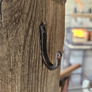 May include: A hand-forged dark metal hook with a curved, tapered end, securely embedded into a vertical piece of weathered, textured brown wood with visible grain patterns and deep vertical fissures running along the surface.