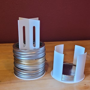 May include: Two white plastic jar lid organizers, one atop a stack of metal jar lids and the other holding a few lids. The organizers have a rectangular shape with vertical slots. The lids are silver and gold-toned, and the background is a wooden surface.