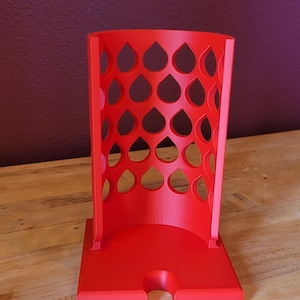 May include: A bright red, 3D-printed phone stand. The stand has a rectangular base with a slot and a cylindrical body with a teardrop pattern cut out. The stand is made of plastic and sits on a wooden surface.