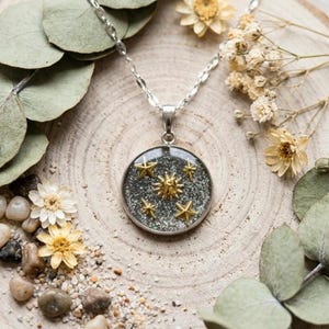 May include: Silver necklace with a circular pendant showcasing gold star details on a glittery base. The necklace is presented on a wooden slice, surrounded by dried flowers and foliage.