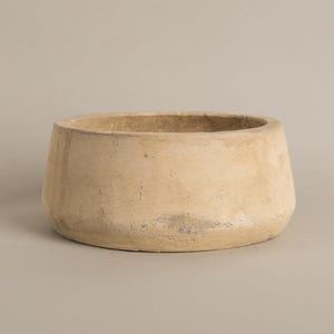 May include: A beige, round ceramic planter with a wide opening and a slightly tapered base. The planter has a textured surface and a neutral colour palette, suitable for various plants and decor styles.