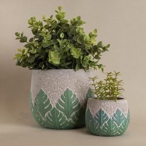 May include: Two decorative plant pots with green foliage. The pots are grey with a textured finish and a green leaf design around the base. The larger pot holds a fuller plant, while the smaller one has a smaller plant.