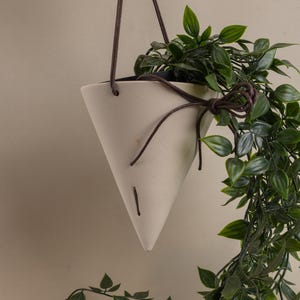 May include: A cream-coloured, cone-shaped hanging planter with a dark brown leather cord. The planter is filled with green leafy plants, with some leaves cascading down. The cord is tied in a decorative knot.