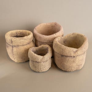 Puede incluir: Cuatro macetas beige, texturizadas, con aspecto de tela, de varios tamaños. Las macetas tienen un aspecto rústico y hecho a mano, con una superficie ligeramente rugosa y un borde superior enrollado. La maceta más pequeña mide unos 8 cm de alto, la más grande unos 15 cm.