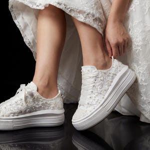 Könnte beinhalten: Weiße Plateau-Sneaker mit aufwendigen Perlen- und Perlenschmuck. Die Sneaker haben weiße Schnürsenkel und eine dicke, weiße Sohle. Die Schuhe sind Teil eines Brautensembles, was auf ein Hochzeitsthema hindeutet.