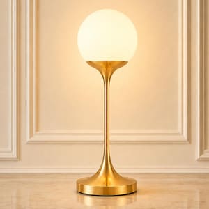 Könnte beinhalten: Eine Tischleuchte mit einem kugelförmigen weißen Glasschirm auf einem schlanken, gebürsteten goldfarbenen Metallschaft und einem runden Fuß. Die Lampe strahlt ein warmes, sanftes Licht aus. Der Text Craftedsoulgiftsshop ist unten rechts sichtbar.