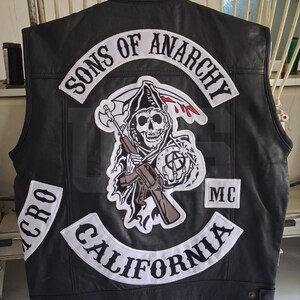 May include: A black leather vest featuring white embroidered patches. The back displays the text SONS OF ANARCHY, CALIFORNIA, and MC. A central patch depicts a skull figure holding a rifle and an anarchy symbol, with red accents.
