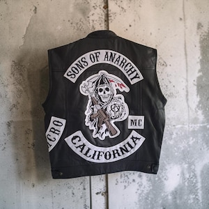 May include: A black leather vest featuring a large embroidered patch on the back. The patch depicts a grim reaper holding a rifle and a scythe, with the text Sons of Anarchy, California, and MC. The vest has a structured collar and button closures.