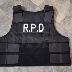 May include: A black tactical vest featuring the white text R.P.D. printed on a rectangular patch across the chest. The vest includes adjustable shoulder straps and two horizontal hook-and-loop fastener strips across the midsection for a secure fit.
