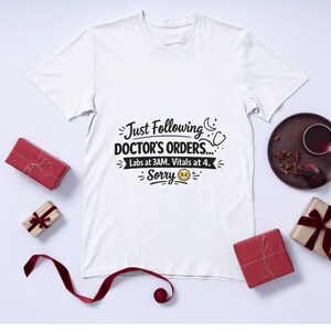 Könnte beinhalten: Weißes T-Shirt mit dem Text "Just Following DOCTOR'S ORDERS... Labs at 3AM. Vitals at 4. Sorry" in Schwarz. Das Shirt ist von verpackten Geschenken und einem dekorativen Teller umgeben.