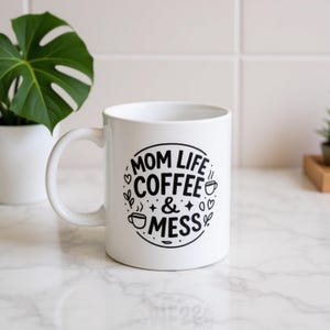 May include: White ceramic mug with the text "MOM LIFE COFFEE & MESS" in a black circular design. The mug has a curved handle and is on a marble surface. A potted plant is in the background.