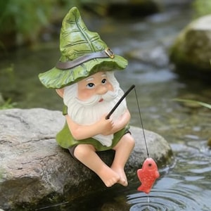 Può includere: Statuina di gnomo da giardino che pesca in un ruscello. Lo gnomo indossa un cappello e un gilet verdi, con barba bianca. Un pesce rosso è all'amo. La scena è ambientata all'aperto, con rocce, acqua e vegetazione.