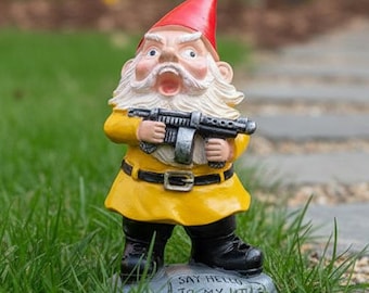 May include: A garden gnome wearing a red hat and yellow coat, holding a machine gun. The gnome has a white beard and stands on a stone base with the text "Say hello to my little friend." The background shows grass and a stone path.