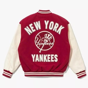 Può includere: Una giacca da baseball rossa e crema dei New York Yankees. La giacca presenta scritte bianche che dicono "NEW YORK" sopra una grafica di baseball e "YANKEES" sotto. Le maniche sono color crema.