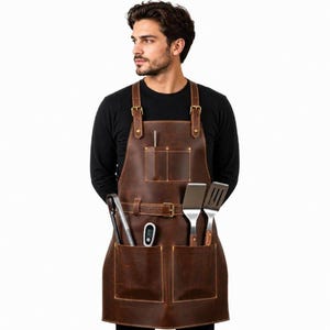 Könnte beinhalten: Eine braune Lederschürze mit verstellbaren Schulterriemen mit Messingschnallen, einer Brusttasche und zwei großen unteren Taschen. Die Schürze verfügt über einen horizontalen Riemen in der Mitte, der Küchenutensilien aus Metall und ein Thermometer hält.