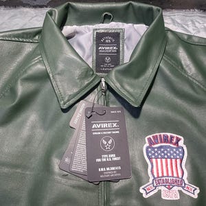 May include: Close-up of a green leather jacket with a collar and zipper. The jacket has a patch with the text "AVIREX ESTABLISHED 1975" and a tag with the text "AVIREX". The jacket is a military-inspired style.