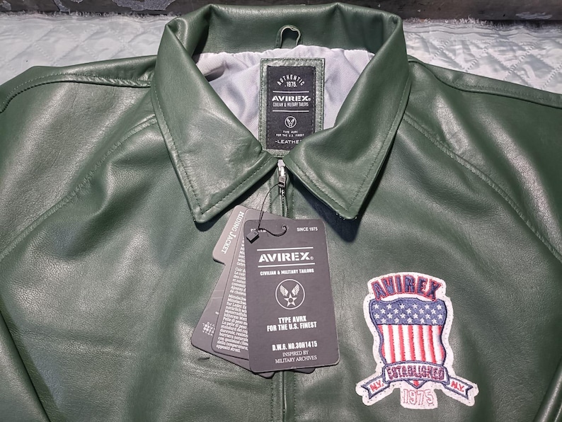 May include: Close-up of a green leather jacket with a collar and zipper. The jacket has a patch with the text "AVIREX ESTABLISHED 1975" and a tag with the text "AVIREX". The jacket is a military-inspired style.