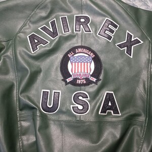 May include: Close-up of the back of a green leather jacket. The jacket features the word "AVIREX" in white block letters across the shoulders, and "USA" below. A circular patch with the American flag is centered.