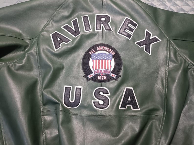 May include: Close-up of the back of a green leather jacket. The jacket features the word "AVIREX" in white block letters across the shoulders, and "USA" below. A circular patch with the American flag is centered.