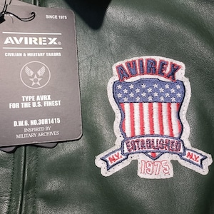May include: Dark green leather jacket with a patch featuring the text "AVIREX" and an American flag design. The jacket has a tag that reads "CIVILIAN & MILITARY TAILORS" and "FOR THE U.S. FINEST". The patch also says "ESTABLISHED 1975".