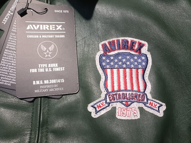 May include: Dark green leather jacket with a patch featuring the text "AVIREX" and an American flag design. The jacket has a tag that reads "CIVILIAN & MILITARY TAILORS" and "FOR THE U.S. FINEST". The patch also says "ESTABLISHED 1975".