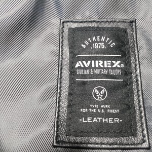 May include: Close-up of a black leather label with white text. The label reads "AUTHENTIC 1975. AVIREX CIVILIAN & MILITARY TAILORS" and "TYPE AURK FOR THE U.S. FINEST -LEATHER-". The label is stitched onto a textured, gray fabric.