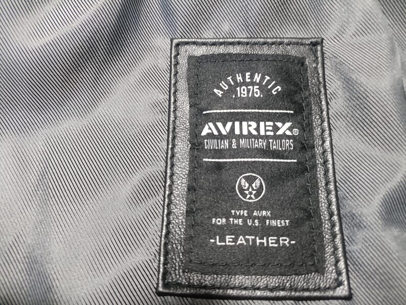 May include: Close-up of a black leather label with white text. The label reads "AUTHENTIC 1975. AVIREX CIVILIAN & MILITARY TAILORS" and "TYPE AURK FOR THE U.S. FINEST -LEATHER-". The label is stitched onto a textured, gray fabric.