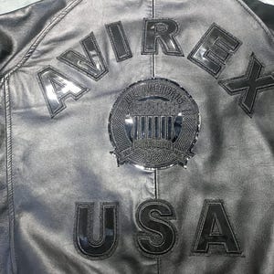 May include: Close-up of a black leather jacket with the word "AVIREX" arched across the upper back. Below the word "AVIREX" is a circular emblem and the word "USA". The jacket has a shiny, reflective finish.